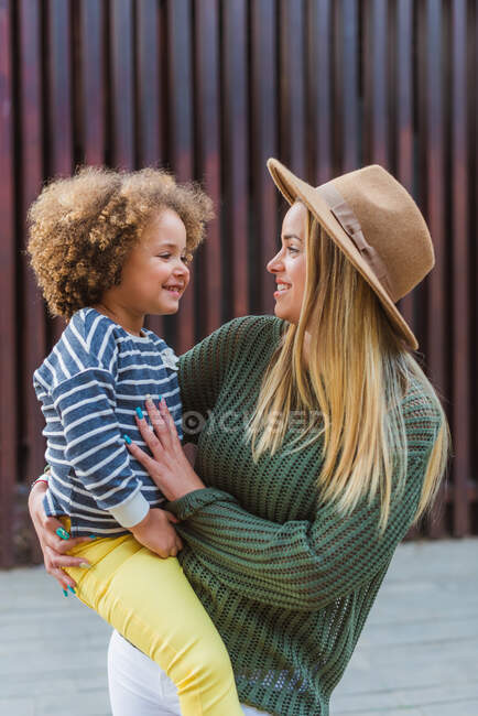 Side view of cheerful young female in trendy sweater and hat embracing cute little daughter while standing on street near modern fence — Stock Photo