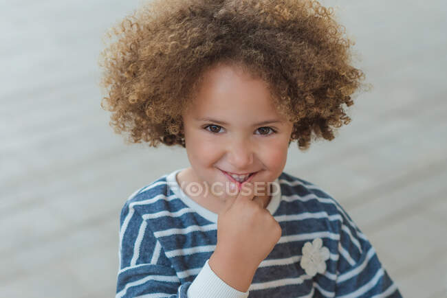 Чарівна маленька дівчинка з кучерявим волоссям в повсякденній смугастій сорочці посміхається дивитися на камеру, стоячи на вулиці — стокове фото