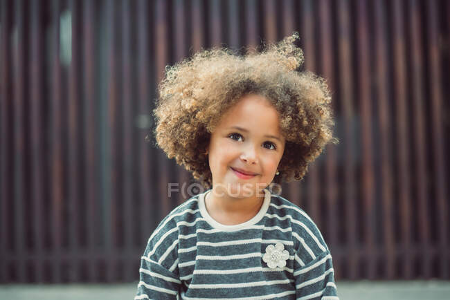 Adorable little girl with curly hair wearing casual striped shirt smiling while standing against blurred wall on street — Stock Photo