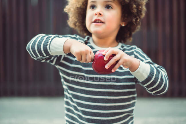 Nettes kleines ethnisches Mädchen mit Afro-Haaren trägt stylisches gestreiftes Hemd, das auf der Straße steht und einen Apfel in der Hand hält — Stockfoto