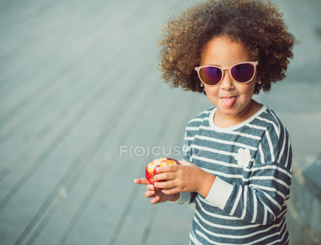 Мила маленька етнічна дівчинка з афро волоссям у стильній смугастій сорочці та сонцезахисних окулярах, посміхаючись та показуючи язик, стоячи на вулиці міста та їдячи яблуко — стокове фото