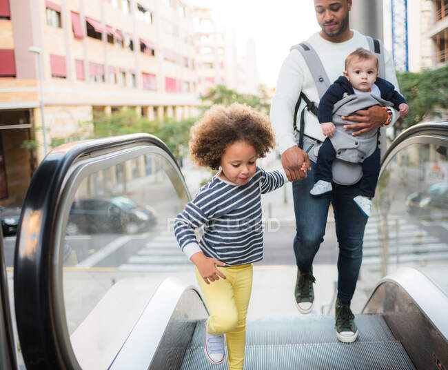 Зверху позитивний молодий етнічний чоловік, що носить немовляти і тримає руку веселої маленької дочки, стоячи на ескалаторі в місті — стокове фото
