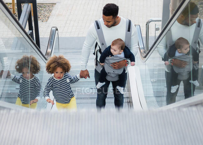 From above of positive young ethnic man carrying infant son and holding hand of cheerful little daughter while standing on escalator in city — Stock Photo