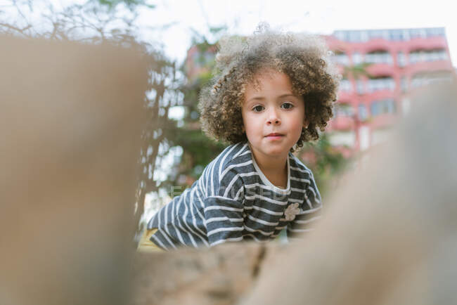 Очаровательная маленькая девочка с вьющимися волосами в повседневной полосатой рубашке, улыбающаяся, глядя в камеру, стоя на улице — стоковое фото
