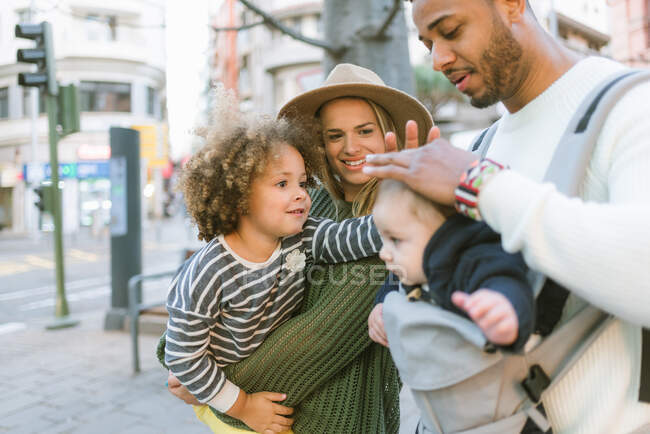 Positif élégant jeunes parents multiraciaux et mignonne petite fille caressant la tête de bébé garçon porté par le père tout en se tenant ensemble dans la rue de la ville — Photo de stock