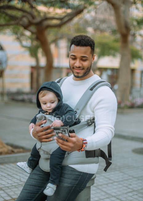 Content African American man embracing son in baby carrier while surfing mobile phone on city street — Stock Photo