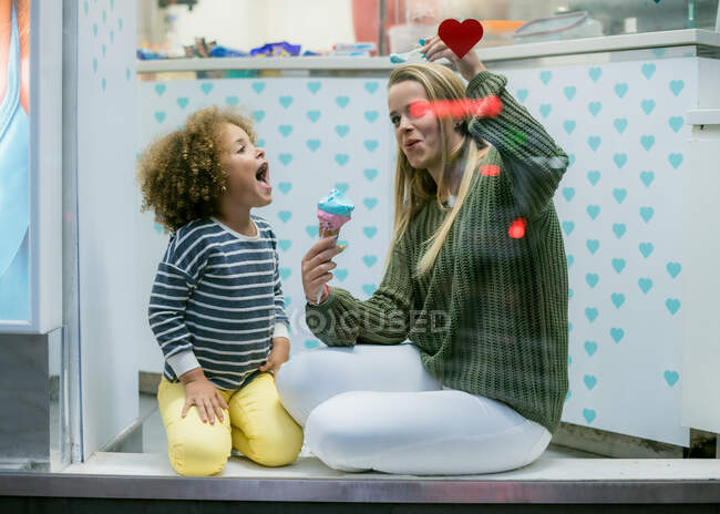 Through cafe window view of loving mother playing airplane with spoon while feeding charming ethnic girl with ice cream — Stock Photo