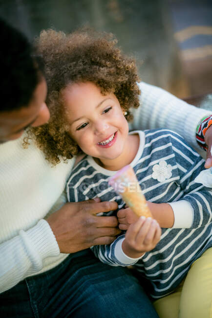 Felice uomo nero guardando affascinante figlia godendo dolce cono gelato mentre riposava sulla panchina in piazza della città — Foto stock