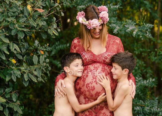 Happy mother in beautiful red dress and floral wreath smiling and embracing twin sons cuddling belly while standing near green trees in garden in summer day — Stock Photo