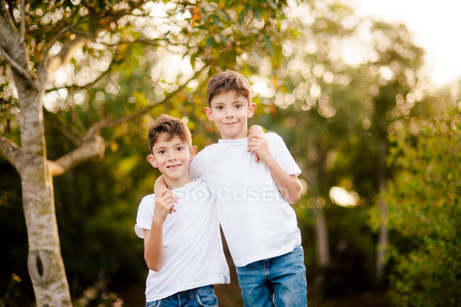 Frères jumeaux souriants en t-shirts et jeans blancs embrassant et regardant la caméra tout en se tenant dans le parc vert dans la journée d'été — Photo de stock
