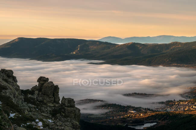 Picturesque drone view of thick white clouds covering valley with town amidst mountains in evening in countryside — Stock Photo