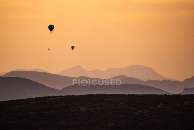 Silhuetas de balões de ar quente distantes voando contra o céu por do sol sobre terreno montanhoso — Fotografia de Stock