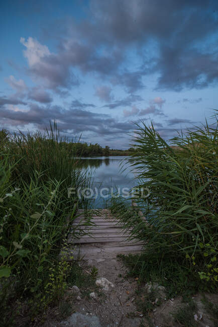 Pequeno cais de madeira localizado na costa gramada da lagoa calma contra o céu nublado de pôr do sol no campo — Fotografia de Stock