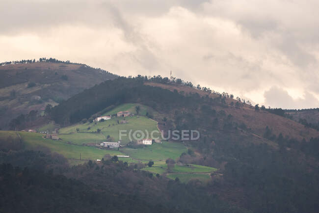 Beaux chalets et champs de ferme situés sur la pente verte de la colline contre un ciel gris et couvert en soirée à la campagne — Photo de stock