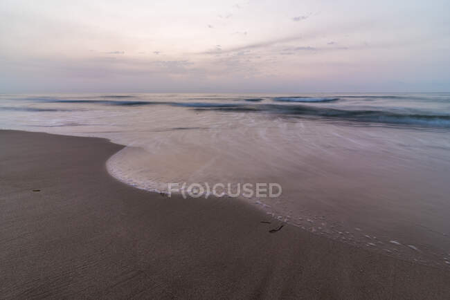 Морские волны катятся по мокрому песчаному побережью против облачного неба вечером в природе — стоковое фото