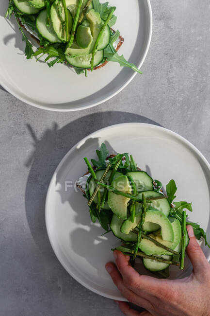 Top view of crop person taking healthy vegetarian toast with green herbs and raw vegetables from white plate — Stock Photo