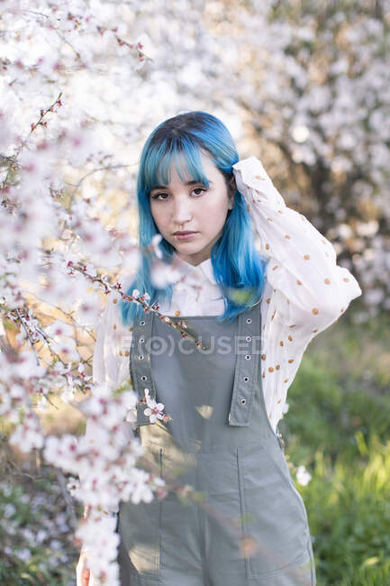 Jovem fêmea elegante com cabelo azul longo olhando para a câmera vestindo elegante geral apreciando árvore florescendo enquanto está em pé no jardim da primavera — Fotografia de Stock