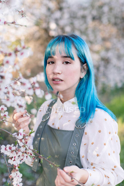 Young stylish female with long blue hair looking at camera wearing trendy overall enjoying blooming tree while standing in spring garden — Stock Photo