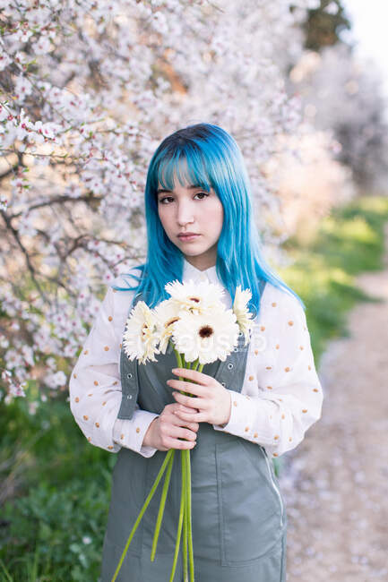 Современная модная женщина с голубыми волосами держит букет свежих цветов и смотрит в камеру, стоя в цветущем весеннем саду — стоковое фото