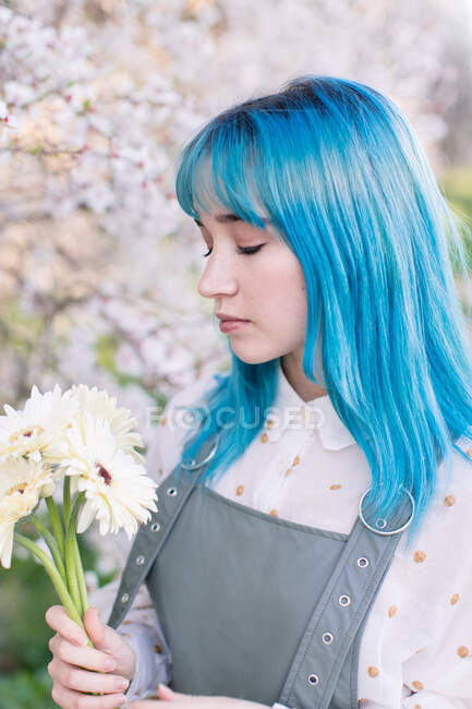 Modern trendy female with blue hair holding and looking at bouquet of fresh flowers while standing in blooming spring garden — Stock Photo