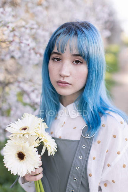 Сучасна модна жінка з блакитним волоссям тримає букет свіжих квітів і дивиться на камеру, стоячи в квітучому весняному саду — стокове фото