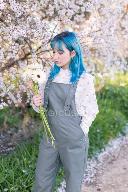 Сучасна модна жінка з блакитним волоссям тримає і дивиться на букет свіжих квітів, стоячи в квітучому весняному саду — стокове фото