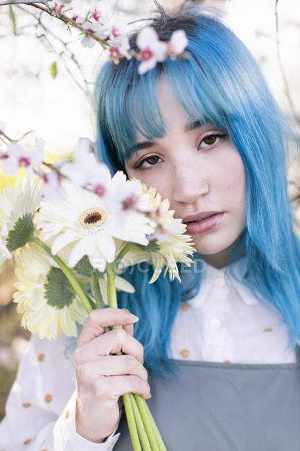 Современная модная женщина с голубыми волосами держит букет свежих цветов и смотрит в камеру, стоя в цветущем весеннем саду — стоковое фото