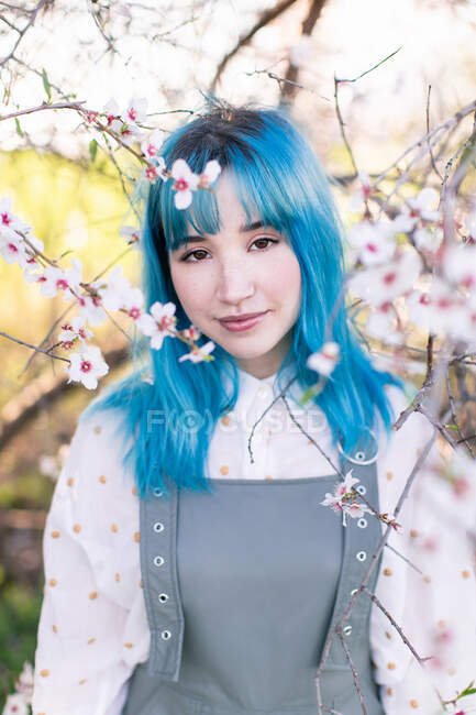 Giovane femmina elegante con lunghi capelli blu guardando la fotocamera indossa trendy generale godendo albero in fiore mentre in piedi in giardino primaverile — Foto stock