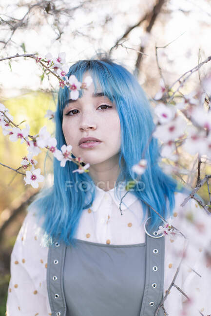 Joven mujer con estilo con el pelo largo y azul mirando a la cámara que lleva moda en general disfrutando del árbol floreciente mientras está de pie en el jardín de primavera - foto de stock
