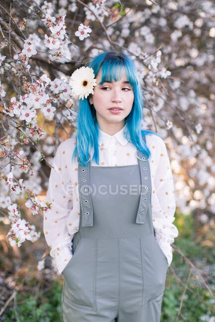 Millennial female model with white flower in blue hair dressed in trendy gray overall standing against blooming sakura tree in garden — Stock Photo