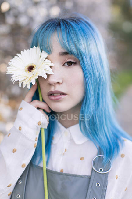 Modernes trendiges Weibchen mit blauem Haar hält ein frisches Blütenauge und blickt in die Kamera, während sie im blühenden Frühlingsgarten steht — Stockfoto