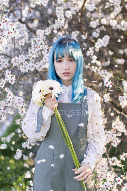 Сучасна модна жінка з блакитним волоссям тримає букет свіжих квітів і дивиться на камеру, стоячи в квітучому весняному саду — стокове фото