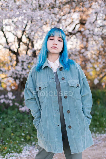 Молодая стильная женщина с длинными голубыми волосами с руками в кармане, смотрящая на камеру, одетая в модный общий вид, наслаждаясь цветущим деревом, стоя в весеннем саду — стоковое фото