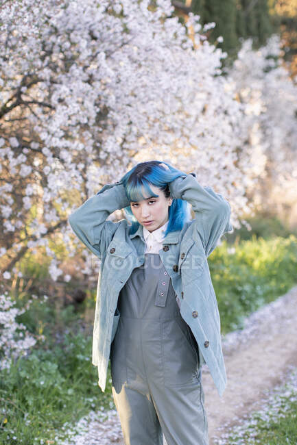 Joven mujer elegante tocando el pelo largo y azul mirando a la cámara usando moda en general disfrutando del árbol floreciente mientras está de pie en el jardín de primavera - foto de stock