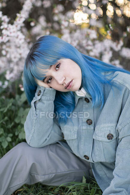 Triste modello femminile millenario con i capelli blu in abito elegante guardando la fotocamera pensieroso mentre seduto su erba verde vicino albero in fiore in giardino primaverile — Foto stock