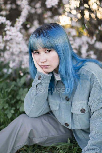 Sad millennial female model with blue hair in stylish outfit looking at camera thoughtfully while sitting on green grass near blooming tree in spring garden — Stock Photo