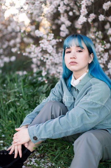 Trauriges Millennial-Model mit blauen Haaren im stylischen Outfit blickt nachdenklich in die Kamera, während sie auf grünem Gras neben blühendem Baum im Frühlingsgarten sitzt — Stockfoto