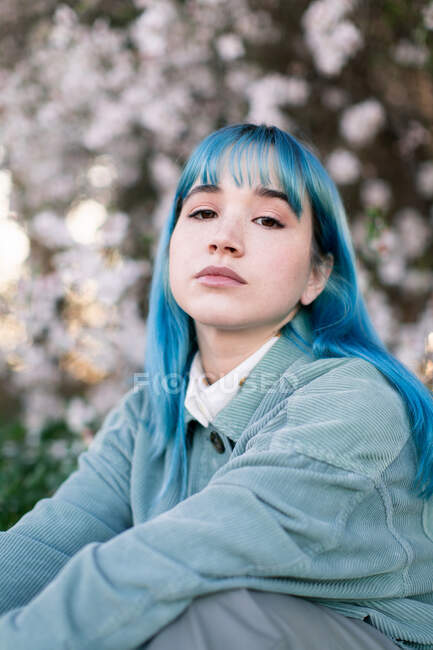 Triste modèle féminin millénaire avec des cheveux bleus dans une tenue élégante en regardant la caméra soigneusement tout en étant assis sur l'herbe verte près de l'arbre en fleurs dans le jardin de printemps — Photo de stock