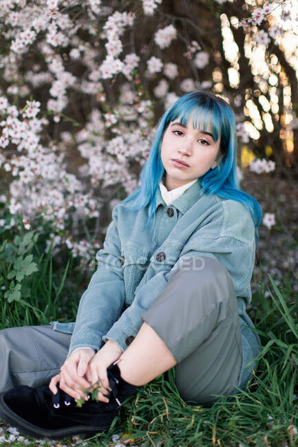 Modelo feminino milenar triste com cabelo azul em roupa elegante olhando para a câmera pensativa enquanto sentado na grama verde perto da árvore florescente no jardim da primavera — Fotografia de Stock