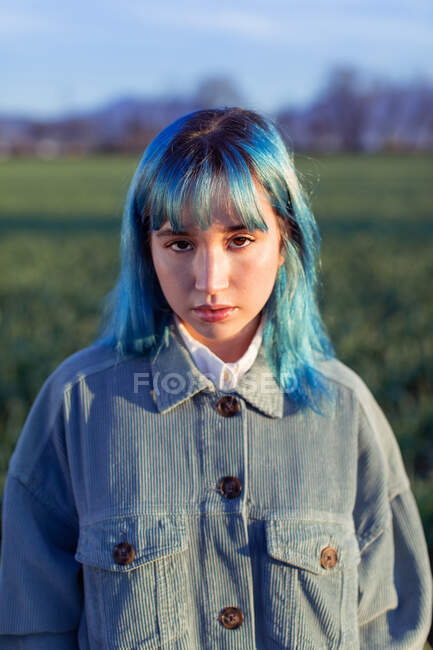 Thoughtful young female with blue hair looking at camera dressed in trendy jacket standing in green field in sunny evening — Stock Photo