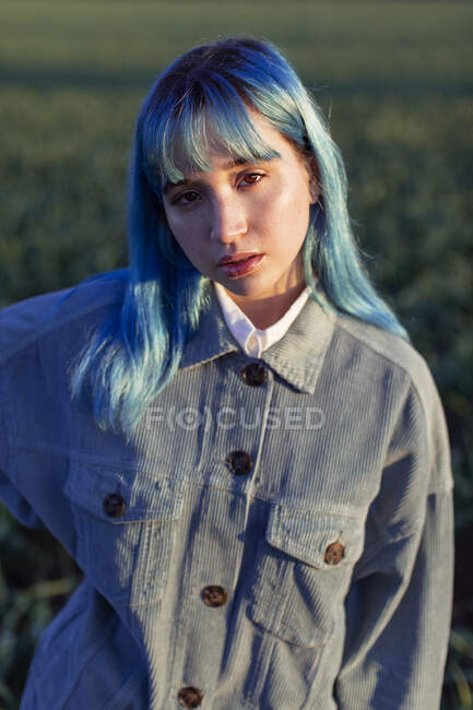 Thoughtful young female with blue hair looking at camera dressed in trendy jacket standing in green field in sunny evening — Stock Photo