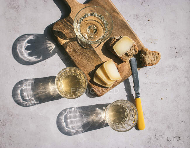 Top view composition with glasses of wine served with sliced cheese and bread with jam on marble table with wooden board in sunlight — Stock Photo