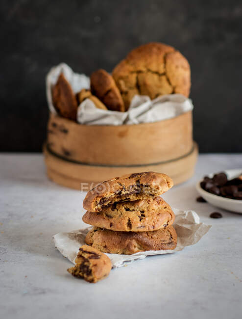 Pila di deliziosi biscotti con gocce di cioccolato con pezzo morso posto sul tavolo vicino a ciotola di legno con pasticceria fresca fatta in casa — Foto stock