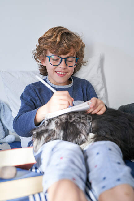 Chico rubio con gafas escribiendo en un cuaderno sentado en la cama con su perro - foto de stock
