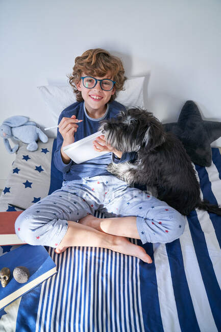 Блондинка в окулярах пише в блокноті, сидячи на ліжку зі своїм собакою — стокове фото