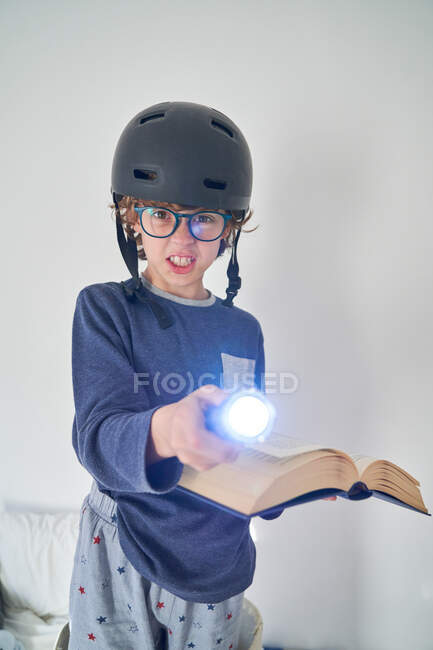 Niño rubio en pijama con un casco una linterna y un libro jugando a la investigación - foto de stock