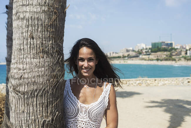 Bella bruna ispanica appoggiata a una palma mentre guarda la fotocamera contro il mare blu — Foto stock
