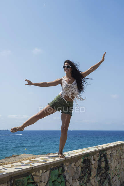 Full-length portrait of carefree brunette woman in shorts dancing on a wall against sea — Stock Photo
