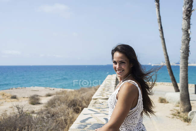 Mulher de cabelos longos em pé à beira-mar enquanto olha para a câmera com um belo sorriso — Fotografia de Stock