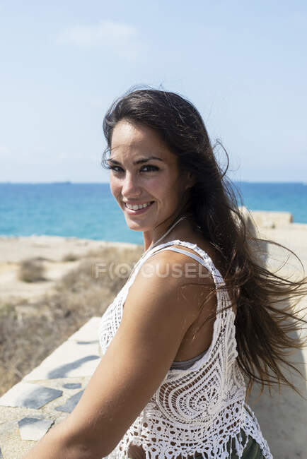 Ziemlich langhaarige Frau, die am Meer steht, während sie mit einem schönen Lächeln in die Kamera schaut — Stockfoto